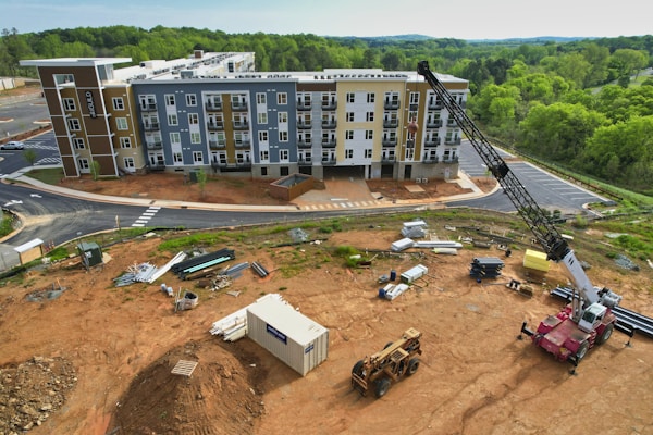 A construction site with a multi-story residential building in the background, surrounded by greenery. Heavy machinery, including a crane and a bulldozer, are present along with various construction materials and equipment scattered across the dirt-covered ground. A container is also visible on the site.
