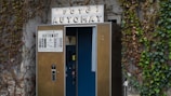 A vintage photo booth with a sign reading 'FOTO AUTOMAT' is situated against a stone wall covered with climbing ivy. The booth has a brown exterior with a slightly open blue curtain revealing a glimpse of the interior, which includes a seat and a photo-taking mechanism. The ivy adds an aged, nostalgic feel to the scene.