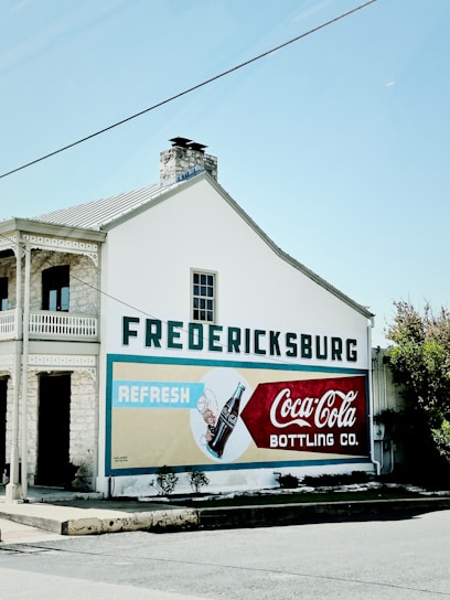 A vintage-style advertisement for Coca-Cola is painted on the side of a building in Fredericksburg. The building has a stone and white facade, with large bold lettering spelling 'Fredericksburg' above the ad. The ad includes imagery of a Coca-Cola bottle and the words 'Refresh' and 'Coca-Cola Bottling Co.'