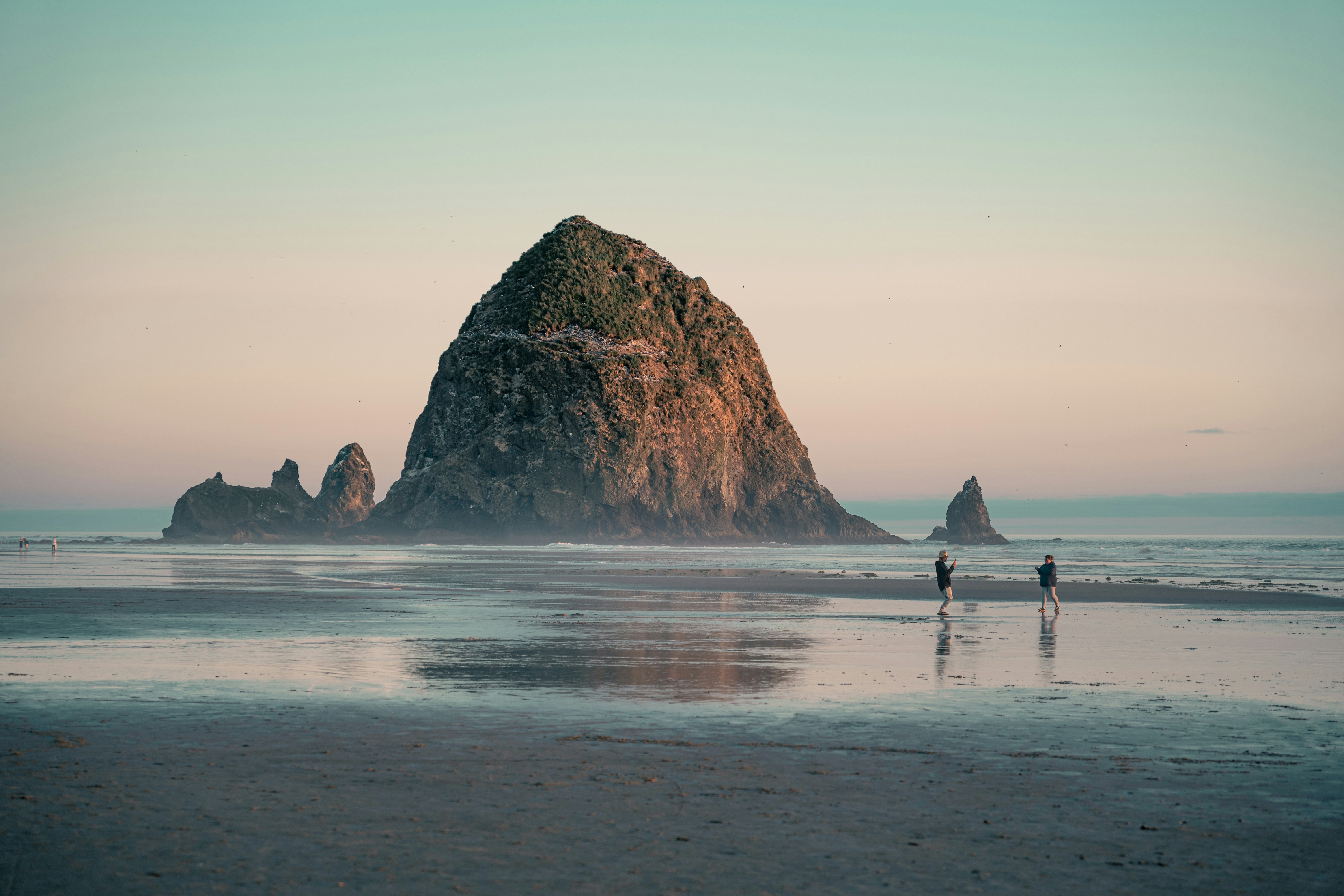 two people standing on a beach next to a large rock