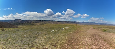 A vast, open landscape with a grassy expanse in the foreground leading to distant mountain ranges. The sky is mostly clear with scattered, fluffy white clouds. A wooden fence marks a trail on the right-hand side, suggesting a natural or hiking setting.