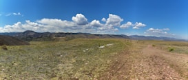 A vast, open landscape with a grassy expanse in the foreground leading to distant mountain ranges. The sky is mostly clear with scattered, fluffy white clouds. A wooden fence marks a trail on the right-hand side, suggesting a natural or hiking setting.
