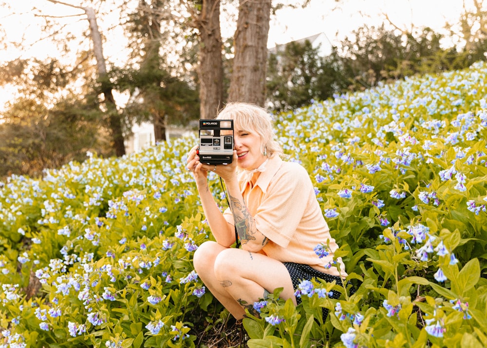 Eine Frau, die in einem Feld mit blauen Blumen kniet und mit einer alten Kamera ein Foto macht