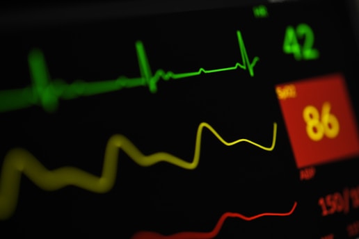 A close-up view of a digital medical monitor displaying vital signs including a green electrocardiogram (ECG) line, a yellow respiratory rate line, and numerical values with a red background indicating a concerning level.