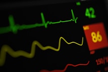 A close-up view of a digital medical monitor displaying vital signs including a green electrocardiogram (ECG) line, a yellow respiratory rate line, and numerical values with a red background indicating a concerning level.