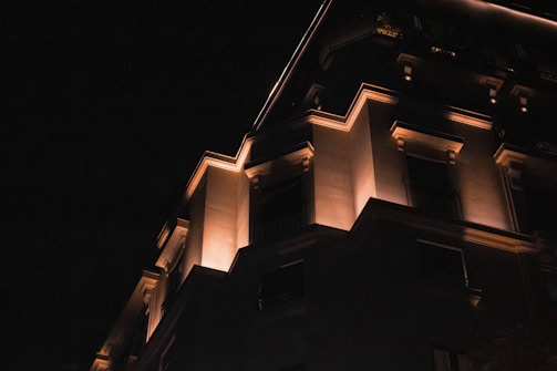 A building facade is illuminated with warm lighting at night, highlighting its architectural features against the dark sky. The structure has a modern design with sharp angles and contrasting shadows.
