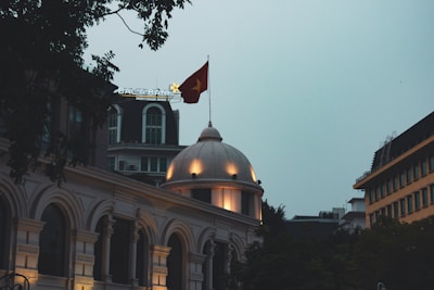 A classical architectural building with arched windows and a central dome displaying a glowing light. Above the dome, a flag with a yellow star on a red background flutters in the dusk sky. In the background, modern buildings and a tree with silhouetted branches contribute to the urban setting.