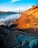 The image depicts a coastal landscape with a prominent orange bridge spanning the background against a bright blue sky. The foreground features a sandy beach with rocks scattered along the shoreline, and steps leading down from the rocky cliffside. Sunlight casts warm hues on the sand and cliffs, enhancing the vibrant colors of the scene.