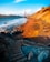 The image depicts a coastal landscape with a prominent orange bridge spanning the background against a bright blue sky. The foreground features a sandy beach with rocks scattered along the shoreline, and steps leading down from the rocky cliffside. Sunlight casts warm hues on the sand and cliffs, enhancing the vibrant colors of the scene.