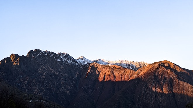 A mountain range is depicted with rugged peaks partially covered in snow. The setting sun casts a warm, golden hue on the ridges, creating a stark contrast with the deep shadows. The sky above is clear and blue, adding to the serene atmosphere.