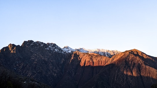 A mountain range is depicted with rugged peaks partially covered in snow. The setting sun casts a warm, golden hue on the ridges, creating a stark contrast with the deep shadows. The sky above is clear and blue, adding to the serene atmosphere.