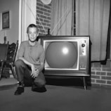 A young boy is kneeling beside a vintage television set with rabbit ear antennas in a living room. The setting includes a brick wall, traditional wooden chairs, and a framed painting on the wall. The image is in black and white, suggesting a mid-20th-century style.