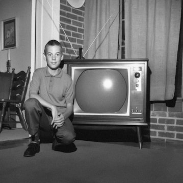 A young boy is kneeling beside a vintage television set with rabbit ear antennas in a living room. The setting includes a brick wall, traditional wooden chairs, and a framed painting on the wall. The image is in black and white, suggesting a mid-20th-century style.