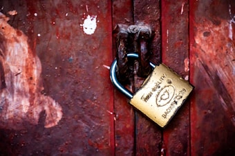 A weathered metal padlock secures the chain on a rusty, red metal door. The lock is inscribed with text and symbols, indicating its high quality. The surface of the door displays various textures and peeling paint, adding an aged appearance.