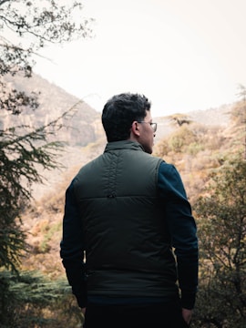 A person wearing a green vest and dark pants is standing outdoors looking at a scenic view of mountains and trees. The person is seen from behind and is wearing glasses. The surrounding environment is natural, with various trees and mountainous terrain.