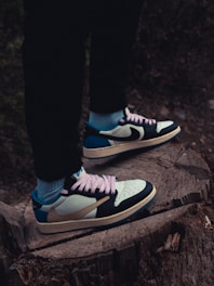 A pair of stylish sneakers with black, white, blue, and tan colors, featuring pink laces. The shoes are worn with blue socks and black pants, standing on a large tree stump outdoors.