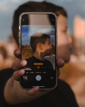 A person is holding a smartphone in front of their face, capturing a side profile image on the screen. The phone is in focus, displaying the camera app interface, with the background slightly blurred.