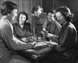 Five women are gathered around a table, engaged in a card game. Some are looking at their cards intently, while others are smiling and interacting with each other. The setting suggests a social and relaxed atmosphere.