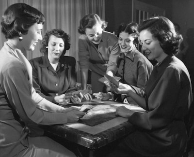 Five women are gathered around a table, engaged in a card game. Some are looking at their cards intently, while others are smiling and interacting with each other. The setting suggests a social and relaxed atmosphere.