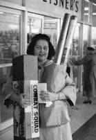 A woman is standing in front of a store, holding several shopping items, including a wrapped gift and long rolls of paper. She appears to be smiling. The store name 'EISNER'S' is visible in the background. Another person is seen in the background, entering or exiting the store.