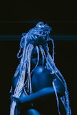 A person with long, intricate braids sits against a dark backdrop, illuminated by blue lighting that casts shadows, creating a dramatic and artistic effect.