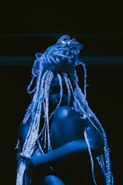 A person with long, intricate braids sits against a dark backdrop, illuminated by blue lighting that casts shadows, creating a dramatic and artistic effect.