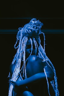 A person with long, intricate braids sits against a dark backdrop, illuminated by blue lighting that casts shadows, creating a dramatic and artistic effect.