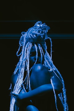 A person with long, intricate braids sits against a dark backdrop, illuminated by blue lighting that casts shadows, creating a dramatic and artistic effect.