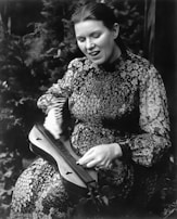 A person is playing a stringed musical instrument, possibly a dulcimer, with a focused and expressive facial expression. The individual is wearing a patterned dress and is captured in a natural, outdoor setting with foliage in the background.