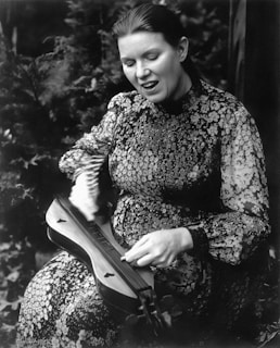 A person is playing a stringed musical instrument, possibly a dulcimer, with a focused and expressive facial expression. The individual is wearing a patterned dress and is captured in a natural, outdoor setting with foliage in the background.