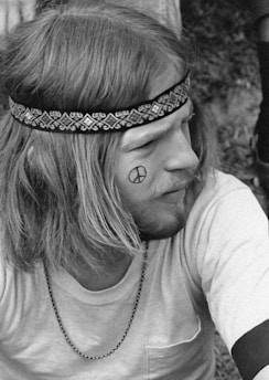 A person with long hair wears a patterned headband and a simple shirt. A peace symbol is drawn on their cheek. They have facial hair and are seen in a relaxed posture outdoors.