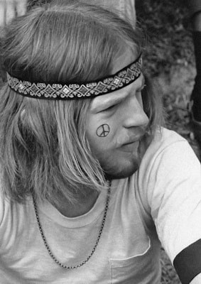 A person with long hair wears a patterned headband and a simple shirt. A peace symbol is drawn on their cheek. They have facial hair and are seen in a relaxed posture outdoors.