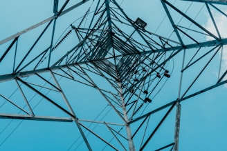 A tall metal structure is seen from below, forming geometric patterns as the perspective narrows upwards. The structure appears to be part of a power transmission tower against a clear blue sky. The intricate lattice of metal beams and electrical components are prominently visible.