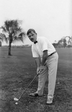 A man is on a golf course, preparing to hit a golf ball with a golf club. He is dressed in a white shirt with a bowtie and light-colored pants. The grass is sparse, and there is a palm tree in the background under an overcast sky.