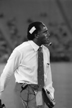 A man is dressed in a white shirt and tie, with a bandage on his head, holding a clipboard and wearing a headset. He appears to be focused and attentive, possibly involved in an event or activity where communication is crucial.