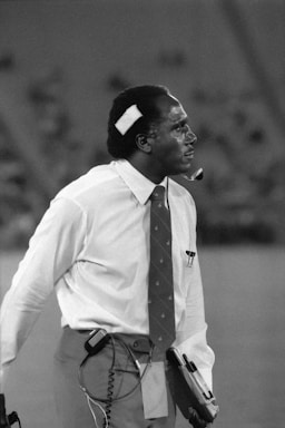 A man is dressed in a white shirt and tie, with a bandage on his head, holding a clipboard and wearing a headset. He appears to be focused and attentive, possibly involved in an event or activity where communication is crucial.