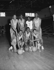 A group of seven young men in athletic uniforms poses with basketballs on an indoor court. They appear to be a part of a basketball team, and each is wearing a numbered jersey. The setting is a gymnasium with bleachers visible in the background, and the lighting suggests a dimly lit space, possibly during a break in a game. One person is holding a trophy in the background.