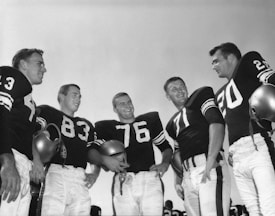 A group of five men standing outdoors in football uniforms, smiling and holding helmets. The men are wearing dark jerseys with large white numbers and light-colored pants. The sky is clear and can be seen in the background.