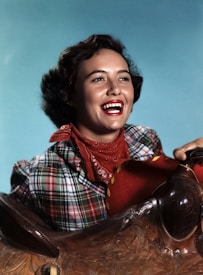 A woman with a joyful expression, wearing a plaid shirt and a red bandana around her neck, leans against an intricately carved saddle. Her dark hair is styled in loose curls, and the background is a solid light blue color.