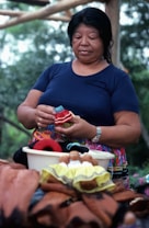 A woman is engaged in crafting, holding a small handmade doll or textile item. She appears focused and calm, surrounded by other colorful objects and fabrics on a table. The background is an outdoor setting with trees, suggesting a natural environment.
