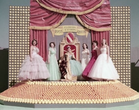 A group of five women dressed in elegant, colorful gowns stand on a stage adorned with numerous citrus fruits. In the center, a woman is seated on a throne-like chair, wearing a royal robe and crown. The backdrop of the stage features a red curtain with gold trim and a sign reading 'Florida Citrus Queen 1955'.