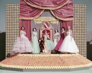 A group of five women dressed in elegant, colorful gowns stand on a stage adorned with numerous citrus fruits. In the center, a woman is seated on a throne-like chair, wearing a royal robe and crown. The backdrop of the stage features a red curtain with gold trim and a sign reading 'Florida Citrus Queen 1955'.