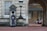 A guard dressed in a formal uniform stands at attention in a sentry box outside a grand historic building with stone walls and large windows. An archway to the right shows part of a courtyard with parked cars and a couple of people walking in the background.