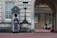 A guard dressed in a formal uniform stands at attention in a sentry box outside a grand historic building with stone walls and large windows. An archway to the right shows part of a courtyard with parked cars and a couple of people walking in the background.