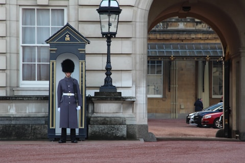 A guard dressed in a formal uniform stands at attention in a sentry box outside a grand historic building with stone walls and large windows. An archway to the right shows part of a courtyard with parked cars and a couple of people walking in the background.