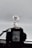 A model or figure of a robot stands prominently, holding a long, thin weapon. The robot features intricate detailing with a color scheme primarily of white, blue, and red, and has distinct yellow accents on its head. It is positioned in front of a camera, which is partially visible and out of focus in the foreground, capturing the figure against a plain, light gray background.