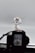 A model or figure of a robot stands prominently, holding a long, thin weapon. The robot features intricate detailing with a color scheme primarily of white, blue, and red, and has distinct yellow accents on its head. It is positioned in front of a camera, which is partially visible and out of focus in the foreground, capturing the figure against a plain, light gray background.