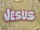 The image features the word 'JESUS' written in large, stylized graffiti letters. The letters are filled with a textured white or light gray color and are outlined with a bold pink or fuchsia color. The graffiti is on a concrete surface that has a weathered appearance and includes a few scattered and faintly chalked markings in various areas.