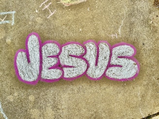 The image features the word 'JESUS' written in large, stylized graffiti letters. The letters are filled with a textured white or light gray color and are outlined with a bold pink or fuchsia color. The graffiti is on a concrete surface that has a weathered appearance and includes a few scattered and faintly chalked markings in various areas.