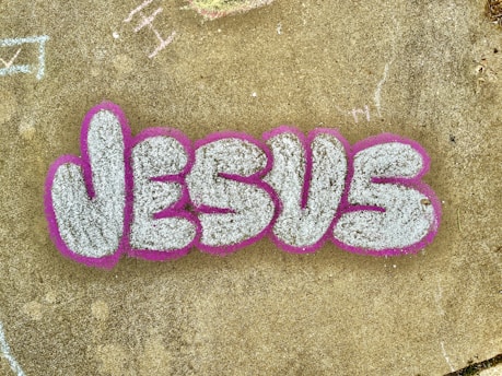 The image features the word 'JESUS' written in large, stylized graffiti letters. The letters are filled with a textured white or light gray color and are outlined with a bold pink or fuchsia color. The graffiti is on a concrete surface that has a weathered appearance and includes a few scattered and faintly chalked markings in various areas.